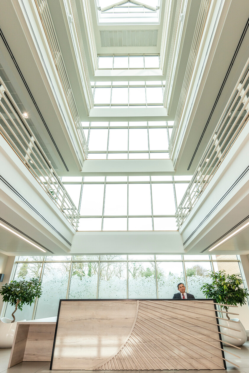 Looking up through a four-storey glazed office atrium to the roof lanterns, with a suited receptionist at the timber desk below