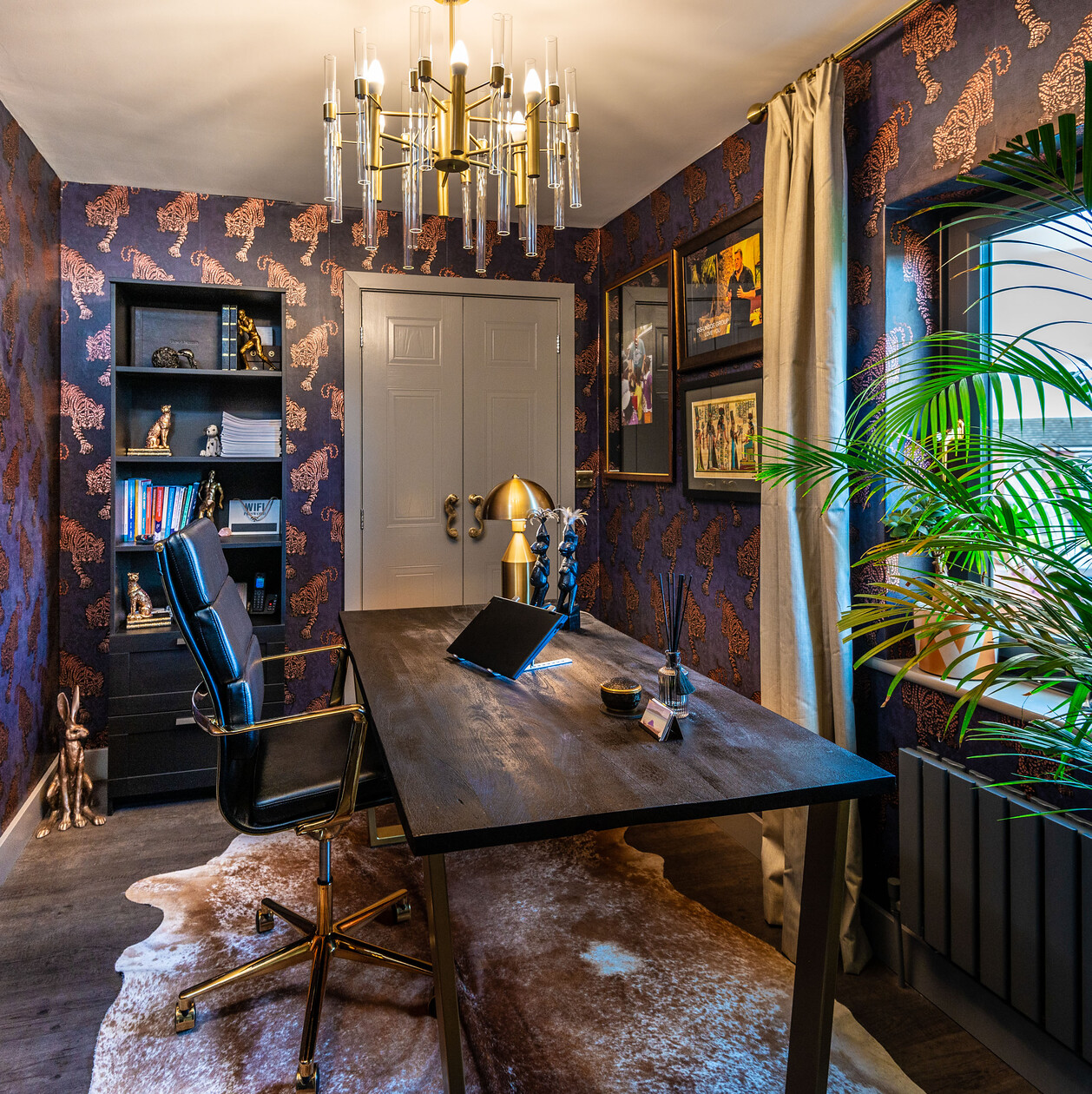 A home study with leopard patterned wallpaper, a modern chandelier, a dark desk, a cowhide rug and framed pictures